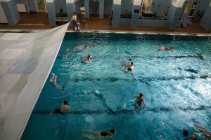 Das halbe Becken eines Hallenbads (Jörgerbad Wien) von oben: ein paar Menschen tummeln sich im Wasser, von links wird eine weiße Plane über das Becken gezogen (Abschlussszene "Swimming Pool", Jörgerbad Wien, © Franzi Kreis)