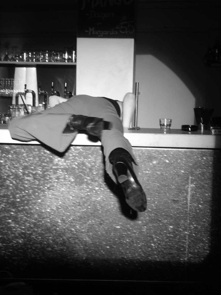 Eszter Hollósi in: "Judit triumphiert": Eine Frau klettert über den Tresen einer Bar, nur die Stiefel mit Absätzen und die Beine in Hosen sind zu sehen, schwarz-weiß.