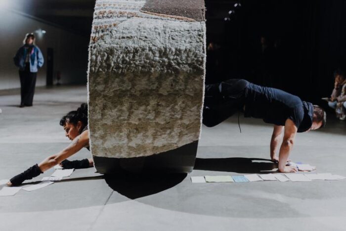 Szene aus der Performance "Roll Over and Over and Over" mit Dafne Moreno und Frederik Maroquin: Dafnes Kopf und Arme ragen aus einem mit Stoff bezogenen Zylinder, Frederik stützt sich mit beiden Händen auf, unter beiden Performenden liegen Kärtchen.